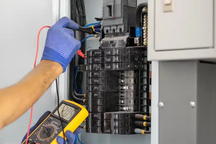 Electrician is using a digital meter to measure the voltage at the circuit breaker control cabinet on the wall.